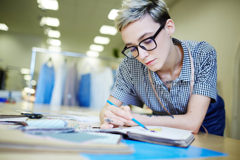 Modedesignerin mit kurzem Haar und Brille in ihrem Atelier und zeichnet Skizzen.