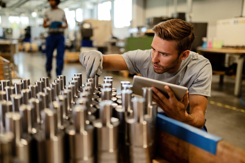 Junger Metallarbeiter mit Touchpad bei der Untersuchung von Stahlstaeben in einer Fabrik vor dem Vertrieb.