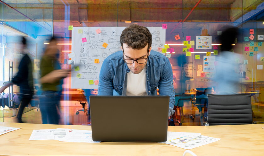 Bild Unternehmenskonzept erstellen Mann mit Brille arbeitet kreativ am Laptop, hinter ihm an Whiteboards vorbeihuschende Menschen.