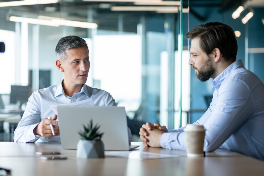 AVGS-Berater diskutiert Businessplan mit maennlichem Gruender in moderner glaeserner Arbeitsumgebung.