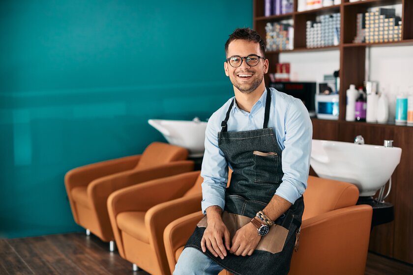 Junger Unternehmer in seinem Friseursalon blickt lächelnd in die Kamera.