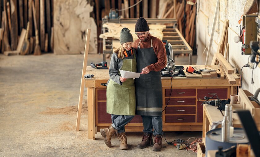 Eine Frau und ein Mann stehen in ihrem Handwerksbetrieb und blicken gemeinsam auf Businessplan.