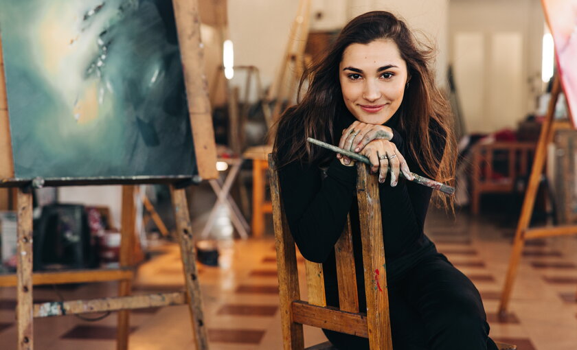 Malerin auf einem Stuhl im Atelier vor ihrer Weinland mit Pinseln in der Hand farbverschmiert gluecklich.