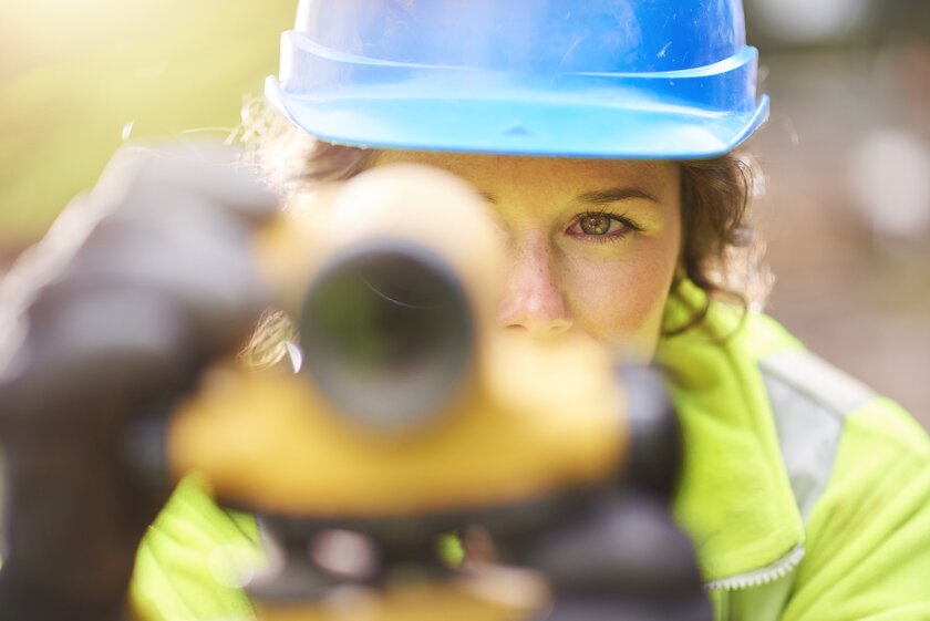 Detailaufnahme einer Frau mit Schutzhelm, die durch eine Vermessungseinrichtung blickt.