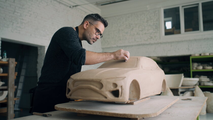 Designer mit Brille und Schuerze arbeitet in einer Keramikwerkstatt an Auto-Prototyp-Skulptur.