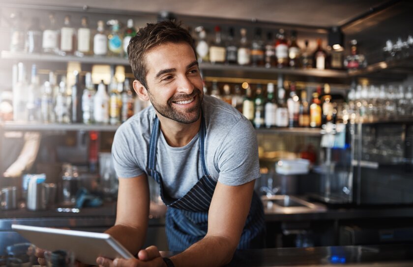 Gastronom arbeitet an digitalem Tablet und blickt freundlich geneigt über Bartresen in den Gastraum.