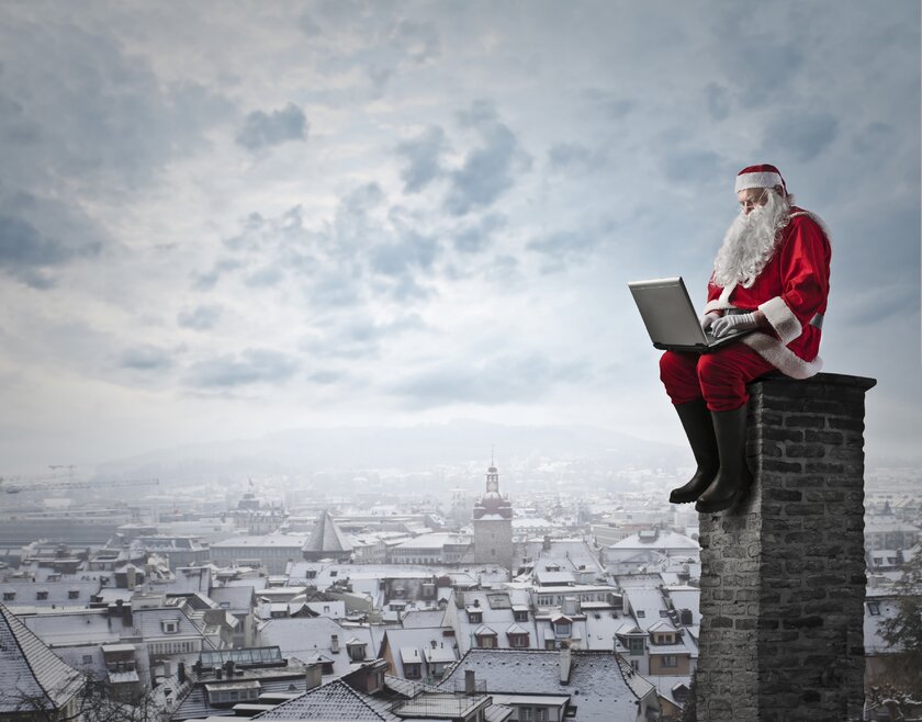Der Weihnachtsmann sitzt mit einem Laptop auf einem Schornstein über einer verschneiten Stadt.