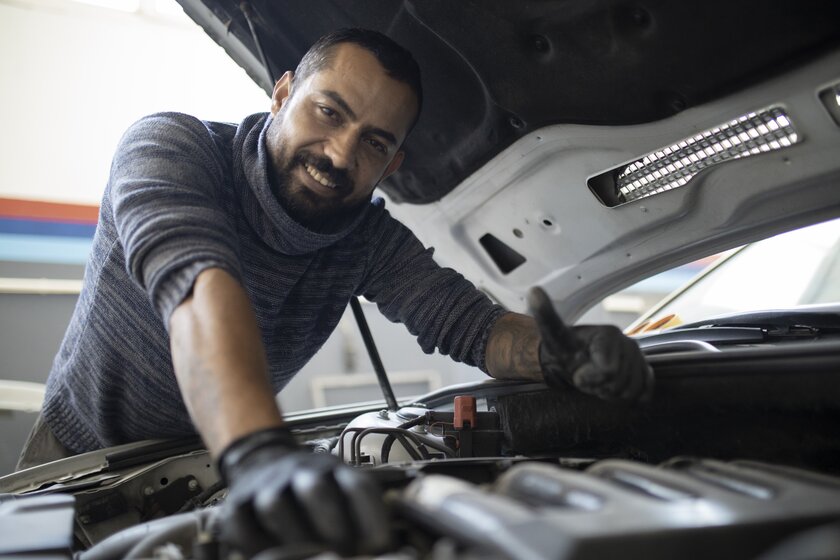 Syrischer KfZ-Mechaniker an seinem Arbeitsplatz einer deutschen Autowerkstatt mit Daumen oben über Motorhaube gebeugt.