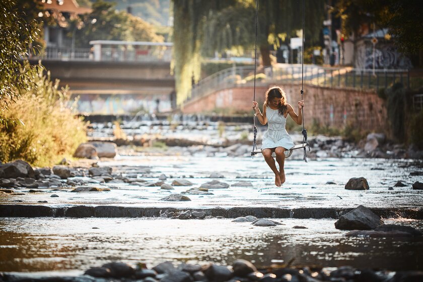 Frau in Sommerkleid sitzt auf einer Schaukel und schwingt über Fluss in Freiburg.