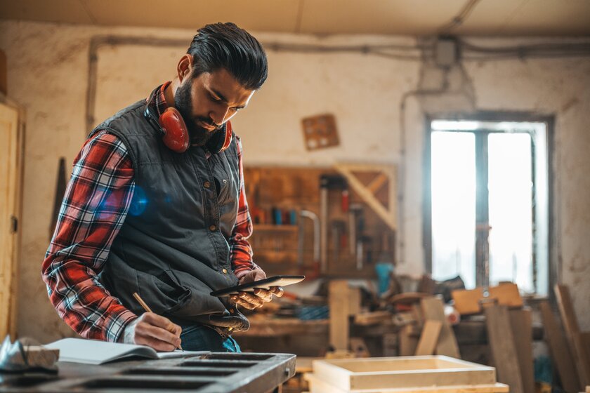 Tischler bei der Arbeit in seiner Werkstatt mit verschiedenen Materialen und Tablet in der Hand.