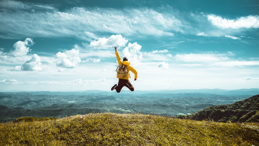 Wanderer mit Rucksack springt auf dem Gipfel eines Berges jubelnd nach oben.