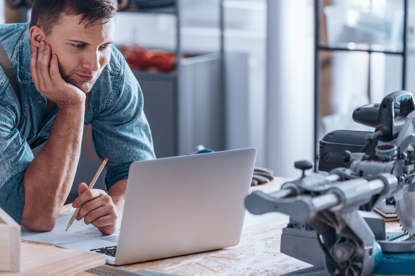 Junger Handwerker in Werkstatt am Laptop denkt nach.