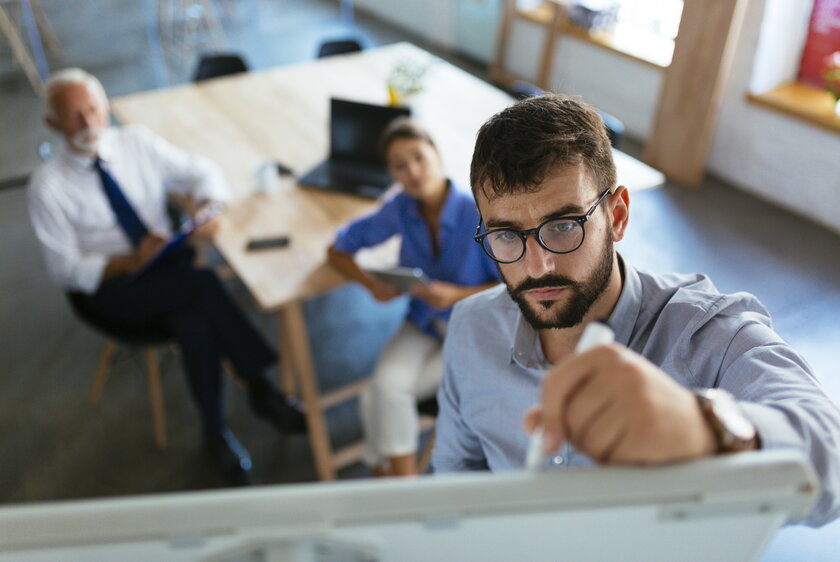 Junger Mann praesentiert Businessplan auf Whiteboard im Konferenzraum vor Investoren.