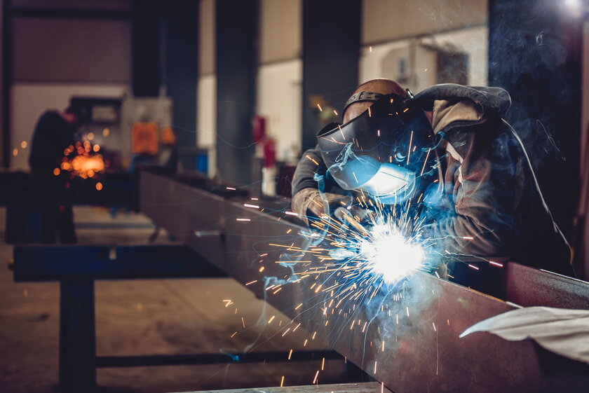  Industrieschweisser mit Funken an Metallwerkstueck in einer Werkshalle.