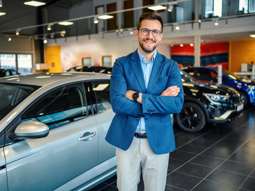 Bild Autohaendler Mann in legerem Business Outfit steht selbstbewusst in Ausstellungsraum vor einer Vielzahl an Autos.