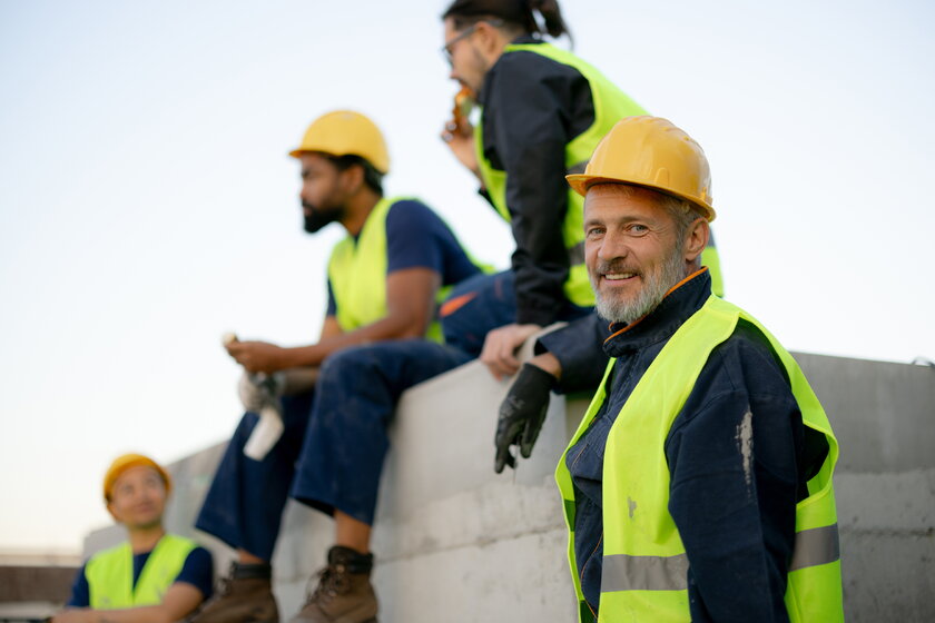 Laechelnder Chef mit Bauarbeitern in Sicherheitsausrüstung, die auf der Baustelle eine Pause einlegen.