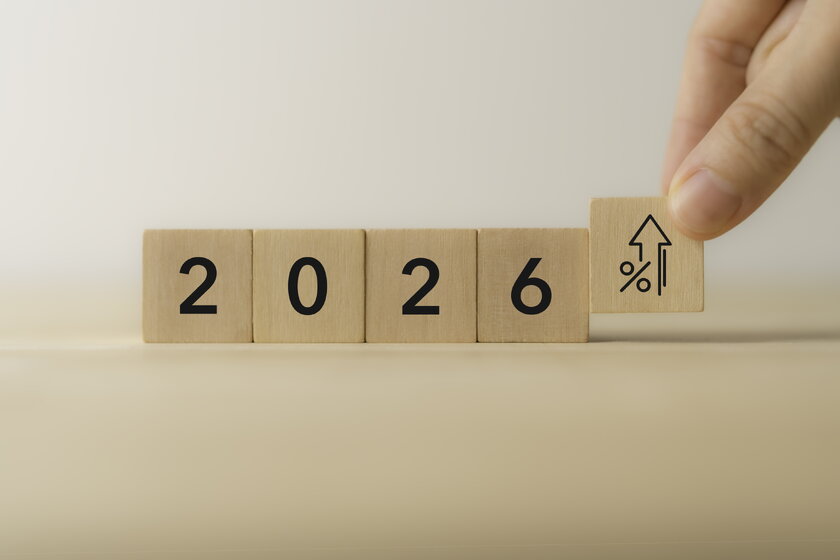 Holzkloetze mit der Aufschrift "2026", eine Hand setzt einen letzten Block mit Aufwaertspfeil und Prozentsymbol.